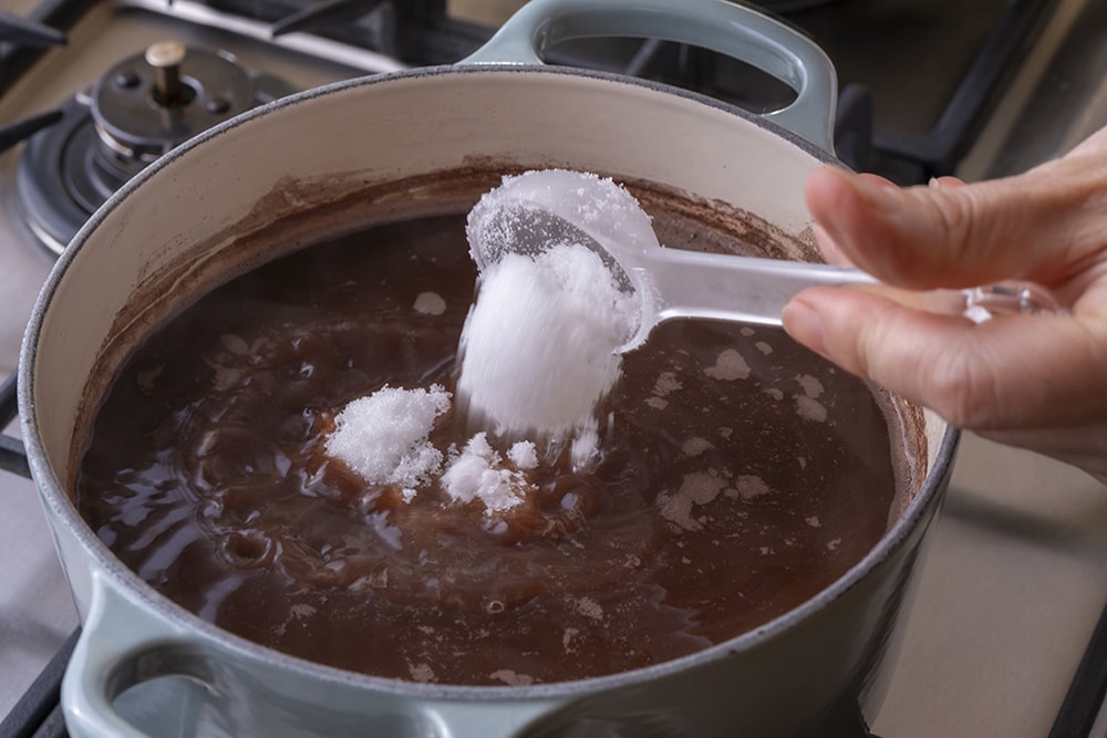 小豆が手で簡単に潰せるくらいまで煮えたら、砂糖を2～3回に分けて入れる。（砂糖を入れるごとに1～2分間煮るとよい。）